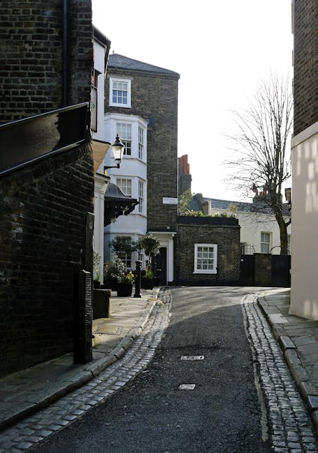 A narrow residential street in Hampstead during daytime, with a cobblestone pavement and curving road. On the left side, there is a dark brick building with a black metal railing and a wall-mounted lantern. In the background, a white bay-windowed house with a small front garden and potted plants can be seen, along with trees that have bare branches indicating winter or early spring. To the right, a white modern building contrasts with the older brick structures. The street appears quiet and is likely used for home relocations and transport of household items. The scene captures the typical setting where professional removals, such as those by Man with Van Hampstead, may operate for local house removals and furniture transport, with the space suitable for loading and unloading moving vehicles.