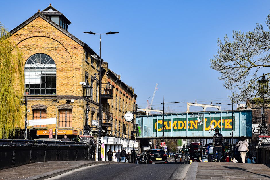 A street scene featuring a large brick building with arched windows and a rounded upper window, situated next to a railway bridge with the yellow and green sign 'Camden Lock' painted on its side. Several pedestrians are walking along the pavement, with some carrying bags or talking, while others wait near parked cars. A man wearing dark clothing is loading or unloading a moving box from a black van, which is parked on the side of the road, close to the building entrance. The scene also includes display signage, street lamps, and trees with budding leaves, indicating a spring day with clear blue sky. This image captures elements relevant to house removals, furniture transport, and packing and moving activities, illustrating the moving process near Camden Lock, with visible materials like cardboard boxes and protective coverings being used during a home relocation facilitated by [COMPANY_NAME], which specialises in removals and moving logistics.