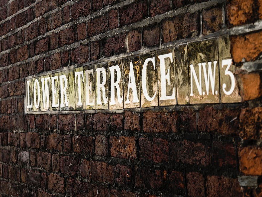 Close-up of a rectangular street sign mounted on a red brick wall, displaying the name 'Lower Terrace' and the postcode 'NW3' in large, white, uppercase letters on a reflective surface. The sign appears slightly weathered with some dirt and moss around the edges, indicating exposure to outdoor elements. The brick wall behind the sign is composed of uneven, reddish-brown bricks with visible mortar. This setting suggests an outdoor residential area, typical of Hampstead, where house removals and furniture transport services like those provided by Man with Van Hampstead may operate. Visible in the background are part of a pavement area, relevant to loading and unloading during home relocation or packing and moving activities. The lighting indicates daylight conditions, emphasizing the textures of the bricks and sign, supporting a professional and factual visual relevant to house removals in Camden.