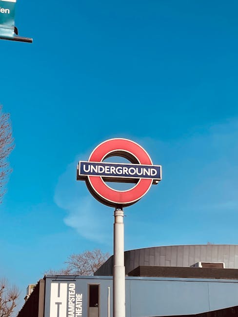 A street scene featuring a large brick building with arched windows and a rounded upper window, situated next to a railway bridge with the yellow and green sign 'Camden Lock' painted on its side. Several pedestrians are walking along the pavement, with some carrying bags or talking, while others wait near parked cars. A man wearing dark clothing is loading or unloading a moving box from a black van, which is parked on the side of the road, close to the building entrance. The scene also includes display signage, street lamps, and trees with budding leaves, indicating a spring day with clear blue sky. This image captures elements relevant to house removals, furniture transport, and packing and moving activities, illustrating the moving process near Camden Lock, with visible materials like cardboard boxes and protective coverings being used during a home relocation facilitated by [COMPANY_NAME], which specialises in removals and moving logistics.