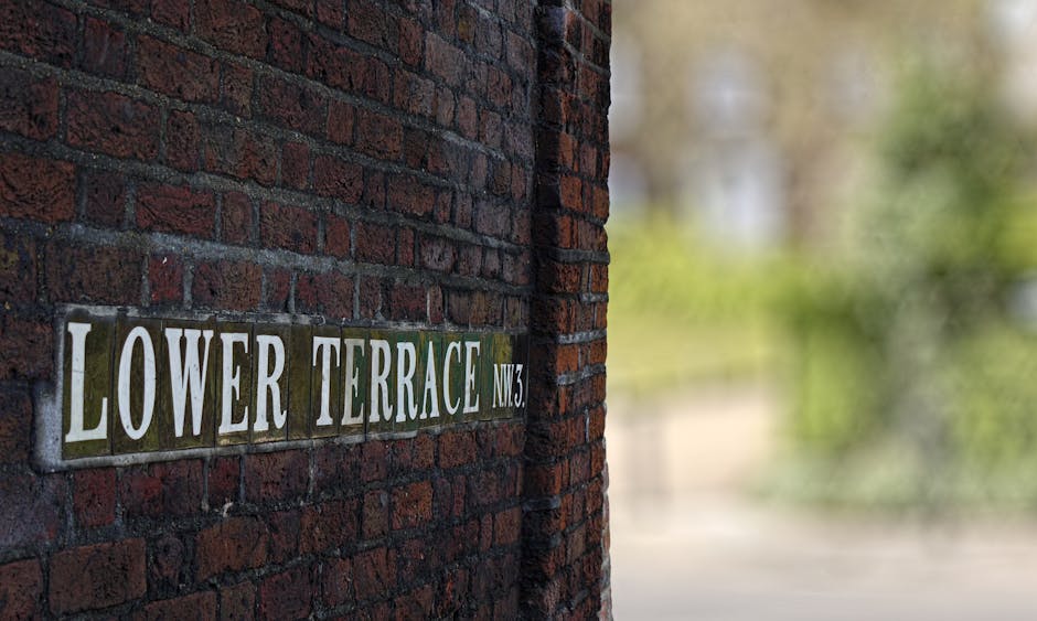 A close-up view of a brick wall displaying a metal house nameplate reading 'LOWER TERRACE NW3' attached to the exterior of a building. The background is blurred, showing greenery and trees, indicating an outdoor setting. This scene is part of a house relocation context, with the focus on the building's address sign that might be relevant during furniture transport or moving logistics for Hampstead homes, as handled by Man with Van Hampstead. The natural light highlights the texture of the bricks and the metal sign, emphasizing the structural elements involved in a typical move or packing process at a property in a narrow access area.