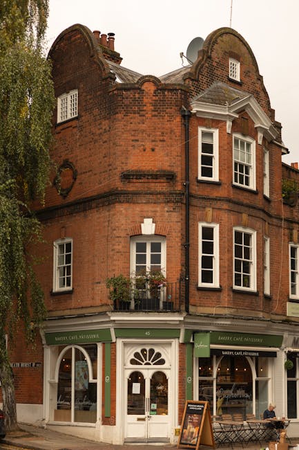 The image depicts a multi-storey red-brick building located on a street corner, with a ground-floor shop featuring a green and white facade, large display windows, and an arched entrance door. The upper floors have several white-framed sash windows, some with small flower boxes and greenery on the window sills, and a decorative balcony with plants. The building's architecture includes curved gables and chimney stacks visible at the roofline. In front of the shop, a person is seated on a bench, and a large tree with leafy branches partly obscures the left side of the building. The scene appears to be taken during daylight hours with natural lighting. The image contextualizes a typical Hampstead street environment, relevant to home relocation or furniture transport services provided by Man with Van Hampstead, illustrating the building's accessibility and the surrounding environment suitable for house removals or moving logistics.