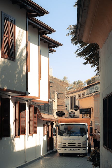A white moving van with the logo of Man with Van Hampstead is parked in a narrow alleyway between two buildings. The scene is outdoors during daylight, with the van positioned close to the buildings' entrances. The building on the left has cream-colored walls, brown wooden shutters on the windows, and a tiled roof extending over the windows. The building on the right has similar architectural features with visible signage for local businesses or shops. Several cardboard boxes and wrapped furniture items are being loaded or unloaded from the van, with some objects positioned near the open rear doors and others on the ground. A person is partially visible near the van, assisting with the loading process. The environment suggests an active home relocation or furniture transport operation, typical of professional moving services provided by companies like Man with Van Hampstead, supporting house removals and packing and moving logistics in the area.