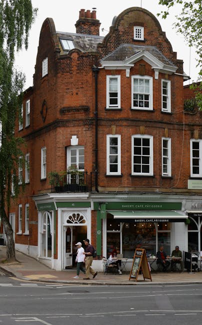 A three-storey red brick building with a rounded gable roof and white-framed sash windows located beside a street. The ground floor features a bakery, café, and patisserie with a green-painted awning and large glass windows. Outside the shop, there are several people walking, some sitting at outdoor tables, and a pedestrian carrying a bag. The pavement is lined with a black metal railing, and the scene is set in daylight with some leafy trees visible on the left side. The image depicts an urban setting typical for house removals and furniture transport in Hampstead, supporting the topic of packing and moving logistics by showing both the building’s exterior and activity on the street.
