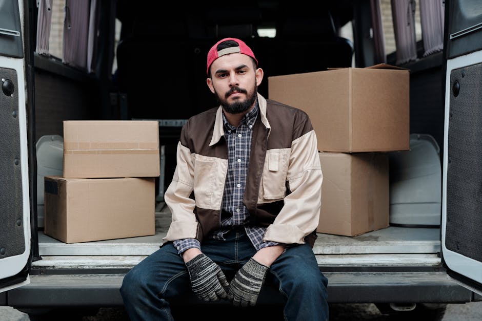 A man with a beard, wearing a red baseball cap backward, a beige and brown jacket over a checked shirt, and dark jeans, is sitting on the edge of an open van's loading platform. Behind him inside the